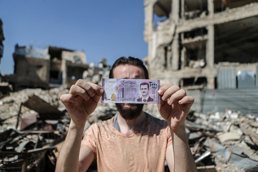 Мъж държи банкнота от 2000 сирийски лири с лика на президента Башар Асад на фона на разрушени сгради в град Доума. Банкнотата бе пусната в обращение в първите дни на месеца